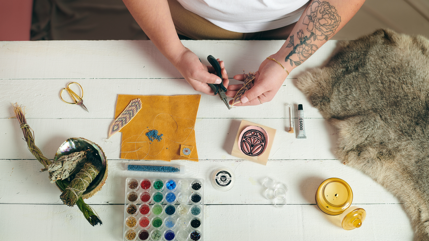 A top-down view of a crafting table where a person is focused on a beading project. The table is filled with colorful beads, tools, and materials, showcasing a creative workspace with a warm, inviting vibe.